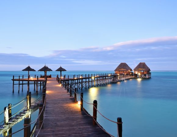 Wooden pier over turquoise water at dusk — thatched over-water lounges and warm lights on the Indian Ocean coast